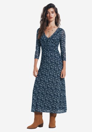 Marineblaues Maxikleid mit Paisley-Muster, V-Ausschnitt, 3/4-langen Spitze-Ärmeln und gerafftem Bund. Kombiniert mit braunen Leder-Stiefeletten.