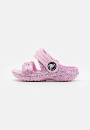 Sandale enfant rose avec détails pailletés, bout ouvert, et bride de talon réglable avec un logo rond noir avec un motif de crocodile.
