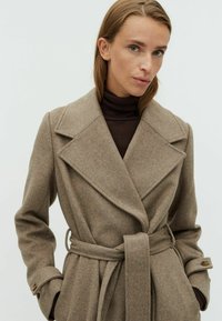 Bruine wolmix jas met een wikkeldesign, grote revers en een stoffen riem. Kenmerkt gebuttonde manchetten en een zachte textuur.