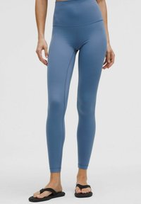 Magas derékszabású, testhezálló leggings puha, rugalmas kék anyagból, sima varrásokkal és látható minták nélkül, flip-flop papuccsal párosítva.