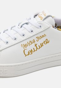 Hvite skinnsko med gullbokstaver "Versace Jeans Couture," med hvite snøringer, rund tå og teksturert gummisåle.