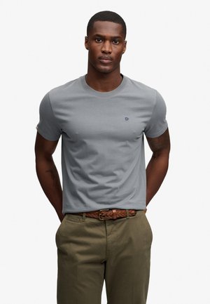 Man standing with hands behind back wearing a gray short-sleeve t-shirt and olive green pants with a braided brown belt.