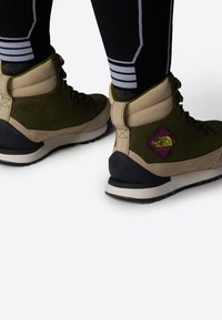 Botas de senderismo verdes y beige con un upper texturizado, suela de goma negra y detalle de logo bordado. Presenta un diseño con cordones y un collar acolchado.