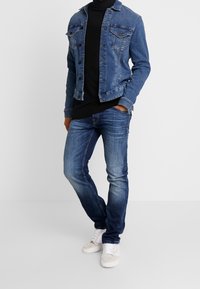Giacca di jeans di blu medio, con due tasche sul petto e bottoni scuri, indossata sopra un maglione a collo alto nero e jeans blu, abbinata a sneakers bianche.