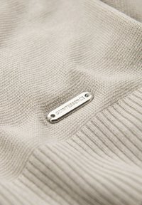 Tejido beige con un patrón de punto texturizado y una etiqueta metálica plateada grabada con "QUINTESSENCE", que presenta dos orificios para sujeción.