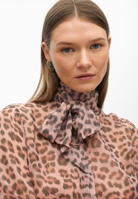 Blusa in stampa leopardata rosa e marrone, con collo alto e fiocco annodato. Materiale leggero con una texture morbida e design fluido.