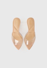 Une paire de sandales beiges à bout pointu, à enfiler, avec de larges brides transparentes et des semelles rembourrées, posées côte à côte sur un fond blanc.