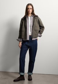 Giacca verde oliva con colletto in velluto a coste marrone, indossata sopra una camicia a quadri e una maglietta bianca, abbinata a jeans di denim scuri e mocassini marroni.