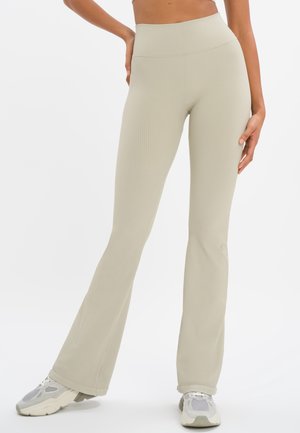 Vrouw draagt beige geribbelde hoge taille flare broek gecombineerd met witte en grijze sneakers, staand met één hand op de heup.