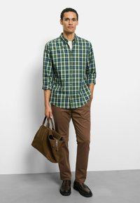 Karierte Hemdjacke in Grün und Blau, braune Hose, braune Tasche und dunkle Schuhe. Das Hemd hat hochgekrempelte Ärmel und abgerundeten Saum.