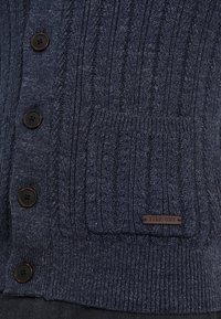 Cardigan lavorato a maglia blu navy con motivi a coste, bottoni marroni, una tasca frontale e un'etichetta in pelle con il logo "PIER-ONE".