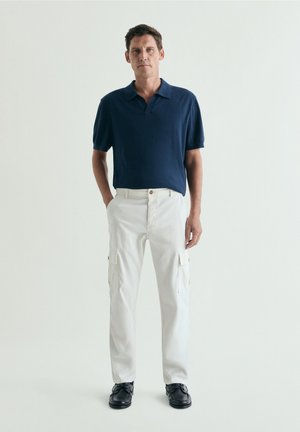 Hombre de pie, vestido con polo azul marino de manga corta, pantalones cargo blancos y zapatos negros, sobre un fondo claro y liso.