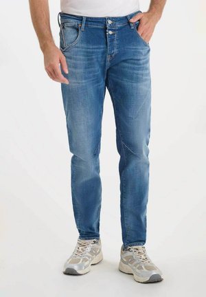 Jean en denim bleu avec une coupe slim, comportant cinq poches, une fermeture à bouton et une fermeture éclair, et un léger délavage sur les jambes. Portés avec des chaussures de sport.