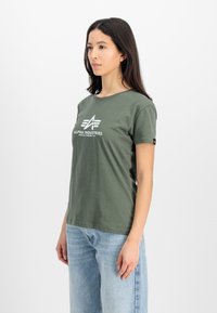 Oliven grøn T-shirt med rund hals, prydet med et hvidt grafisk logo "ALPHA INDUSTRIES" foran og korte ærmer. Lyseblå jeans.
