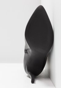 Botte à talon en cuir noir positionnée semelle vers le haut contre un mur blanc en angle, mettant en valeur un bout pointu et un talon aiguille fin.