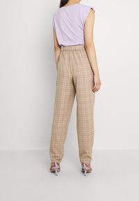 Kvinna står med ryggen mot oss och bär beige rutiga högmidjade byxor, lilafärgad ärmlös topp och matchande lilafärgade klacksandaler.