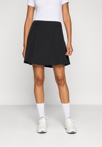 Gonna nera plissettata con vita aderente, realizzata in un materiale liscio. Indossata con una camicia bianca, abbinata a scarpe sportive bianche e calzini.