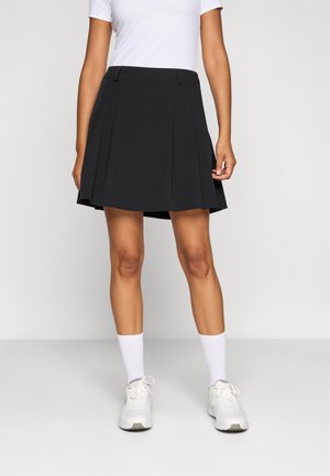 Jupe plissée noire avec une taille cintrée, fabriquée en matériau lisse. Portée avec une chemise blanche, associée à des chaussures de sport blanches et des chaussettes.