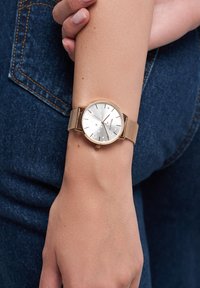 Runder silberner Uhr mit minimalistischer Gestaltung, goldfarbener Mesh-Armband und Sternakzenten auf dem Zifferblatt, auf einer Hand gegen Denim liegend.