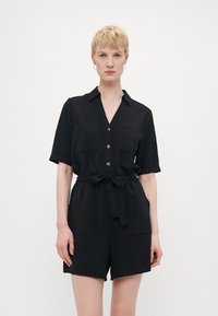 Svart kortermet jumpsuit med krage, frontlukking med knapper, to brystlommer og belte med knyting i midjen. Teksturert stoff.