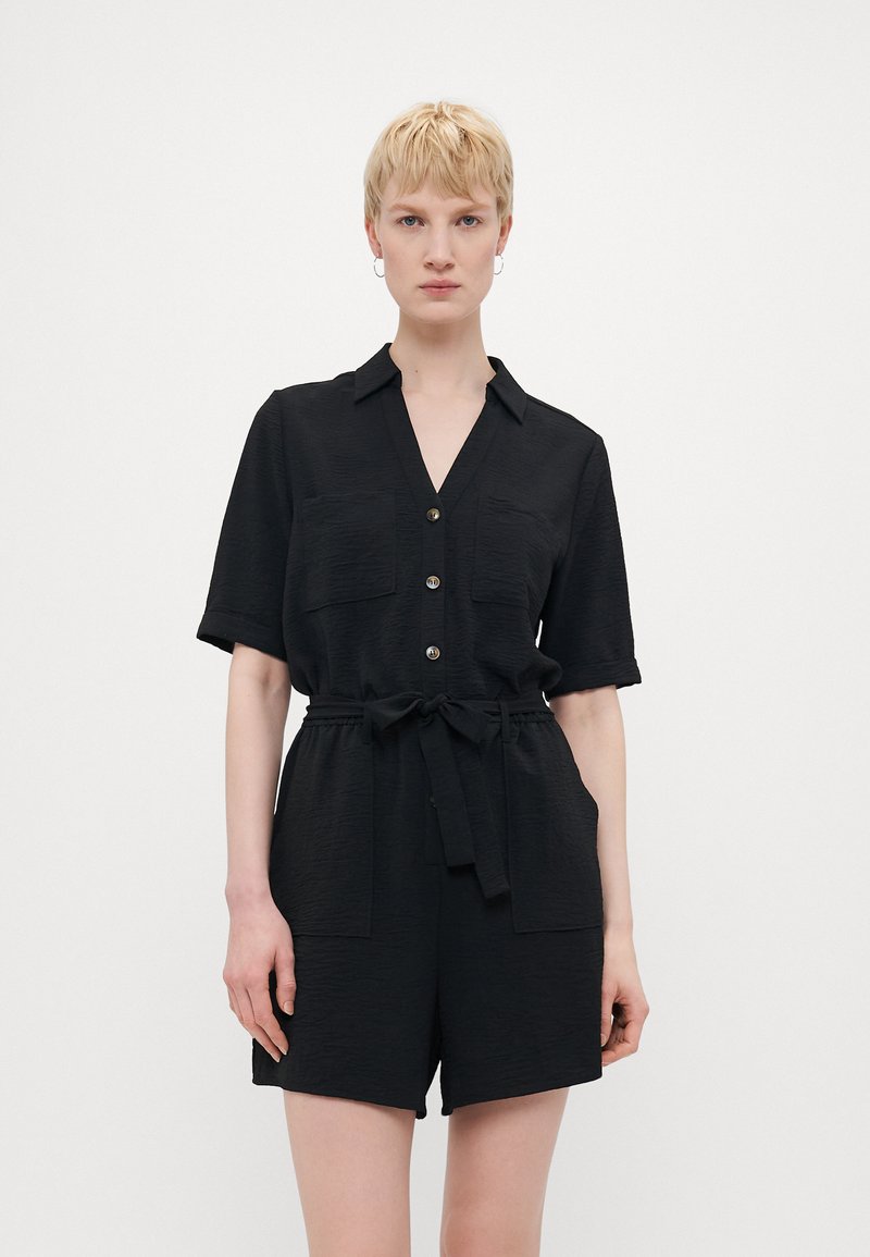 Svart kortermet jumpsuit med krage, frontlukking med knapper, to brystlommer og belte med knyting i midjen. Teksturert stoff.