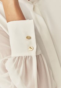 Weißes Blusenoberteil mit transparentem Puffärmel, der mit einem formellen Bündchen verziert ist, das mit zwei glänzenden goldenen Knöpfen geschmückt ist. Weiche, fließende Stofftextur.