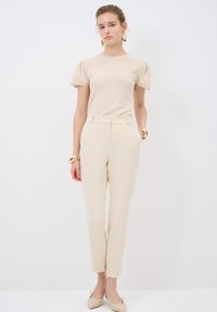 Beige fitted top with puff sleeves, paired with high-waisted beige trousers. Model wears beige pointed flats and gold bracelets. Simple design.