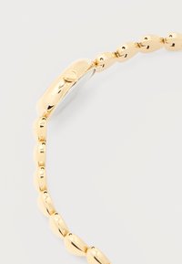 Μεταλλικό μπρασελέ ρολογιού σε χρυσό τόνο με γυαλιστερές, στρογγυλεμένες συνδέσεις και πλάγια όψη του μηχανισμού κλειδώματος πάνω σε λευκό φόντο.