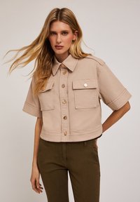 Giacca beige corta a maniche corte realizzata in tessuto morbido, con due tasche frontali, chiusura a bottoni e un design del colletto classico.