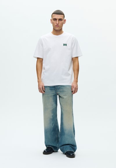 Άσπρο βαμβακερό t-shirt με πράσινο κεντητό λογότυπο, σε συνδυασμό με φαρδιά ξεβαμμένα μπλε τζιν. Το μοντέλο στέκεται σε απλό φόντο. Μαύρα παπούτσια.