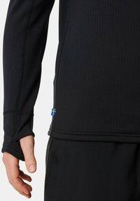 Person trägt ein schwarzes Langarmoberteil mit Daumenloch und einem kleinen blauen Etikett mit einem gelben Kreuz nahe dem Saum.