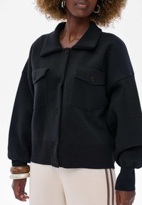 Czarny, oversize sweter z punktowym kołnierzykiem, zapięciem na guziki i dwiema kieszeniami na piersi. Rybowe mankiety i dół wzmacniają teksturowany design.