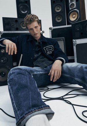 Un jeune homme en veste foncée et jeans est assis parmi de grands haut-parleurs, avec des câbles audio enroulés sur le sol.