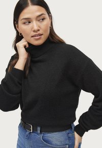 Maglione nero a collo alto con lavorazione a trama, polsini a coste e vestibilità rilassata, abbinato a jeans blu e una cintura nera.