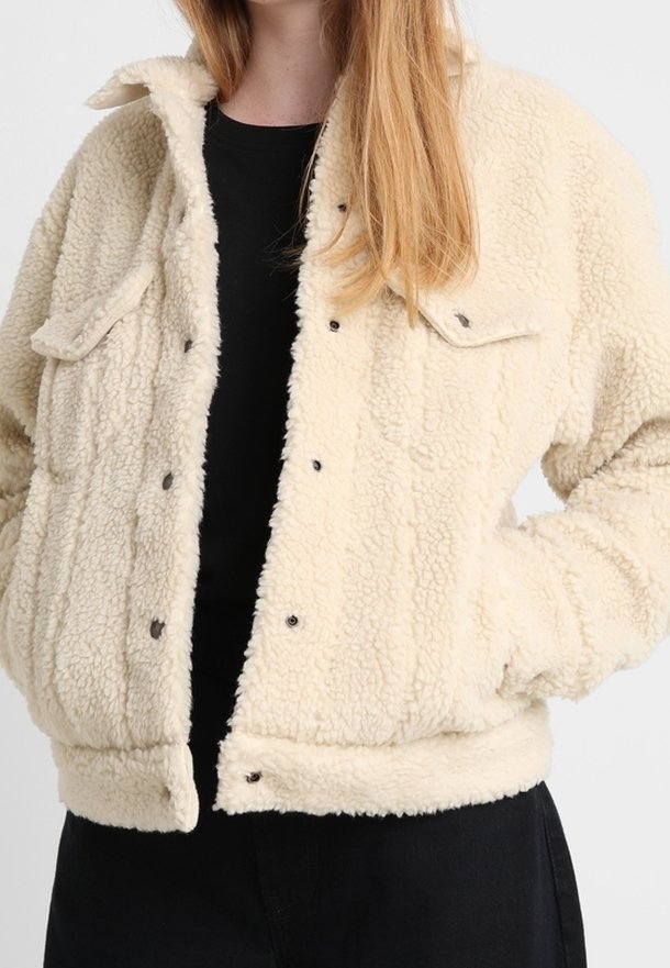 Veste en polaire couleur crème avec une surface texturée, des fermetures à bouton et deux poches avant. Elle présente une coupe décontractée et des manches à poignets.