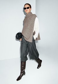 Bruine geribbelde gebreide mouwloze trui, gelaagd over een witte shirt, gecombineerd met grijze wijde broek en glanzende bruine hoge laarzen. Zwarte tas en zonnebril.