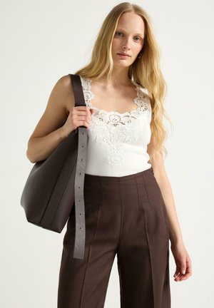 Kvinde med langt blondt hår iført hvid tanktop med blonder, høj taljede brune bukser og bærer en stor mørk brun skuldertaske.