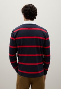 Uomo con i capelli ricci che indossa una camicia a maniche lunghe blu navy con strisce orizzontali rosse e pantaloni beige, di fronte a un muro bianco uniforme.