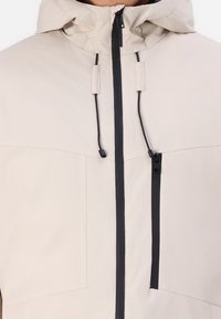 Hellbeige Jacke mit einem Reißverschluss vorne, schwarzen Reißverschlussakzenten, verstellbarem Kordelzugkapuze und zwei Brusttaschen. Glatte Textur.