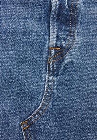 Tissu denim bleu en gros plan avec des surpiqûres visibles jaune-orange formant une partie d'un bord de poche et d'une couture.