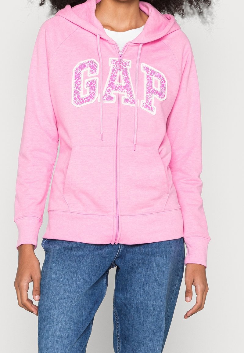 Sudadera rosa con cremallera que presenta un logo de "GAP" con patrones florales, bolsillos tipo canguro y capucha con cordón. Fabricada en un tejido suave y texturizado.