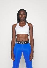 Bela športna nedrčka z črnim logotipom Nike, v kombinaciji z modrimi leginsami, ki imajo črn pas "NIKE PRO". Gladek, oprijet dizajn.