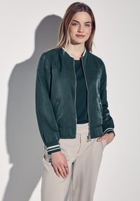 Veste bomber vert foncé avec fermeture éclair à l'avant, poignets et col côtelés. Texture lisse avec un léger brillant ; associée à un pantalon beige clair.