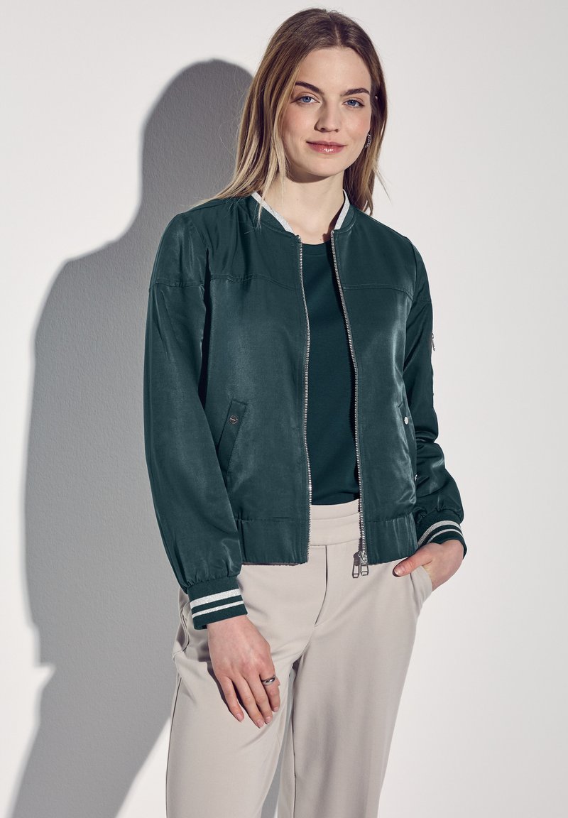 Veste bomber vert foncé avec fermeture éclair à l'avant, poignets et col côtelés. Texture lisse avec un léger brillant ; associée à un pantalon beige clair.
