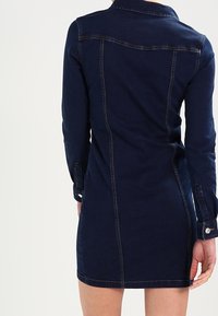 Kvinna som bär en figursydd, långärmad, mörkblå denimklänning med synliga sömdetaljer, visad från baksidan mot en neutral bakgrund.