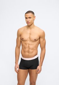 Νεαρός μυώδης άνδρας με κοντό μαλλί, φορώντας μαύρα μποξεράκια με ανοιχτό ροζ μέση, στέκεται μπροστά σε απλό λευκό φόντο.