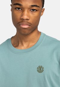 Lyseblå bomulds T-shirt med rund halsudskæring og korte ærmer. Har et mørkegrønt logo på venstre bryst. Glat tekstur.