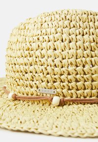 Sombrero de paja en amarillo claro con textura tejida. Presenta un cordón marrón y cuentas de madera blancas, acentuado por una etiqueta plateada de "Roxy".