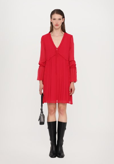 Robe en chiffon rouge avec un col en V, des accents volantés et des manches longues. Portée avec des bottes noires jusqu'aux genoux et tenant un petit sac noir.