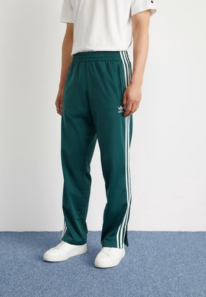 Personne portant un pantalon de survêtement Adidas vert foncé avec des bandes blanches et des baskets blanches, debout sur un tapis bleu contre un mur blanc.